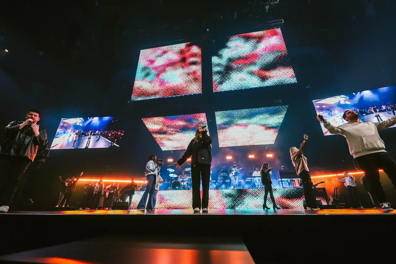 Performers on a dark stage under floating LED screens showing abstract color