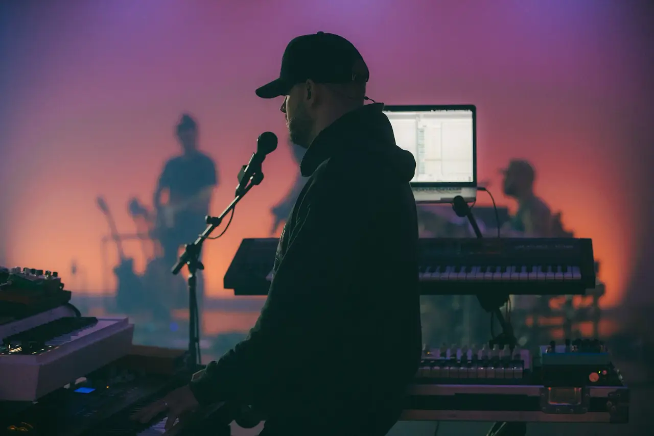 Keyboard player in a cap at the microphone, lit in pink and orange
