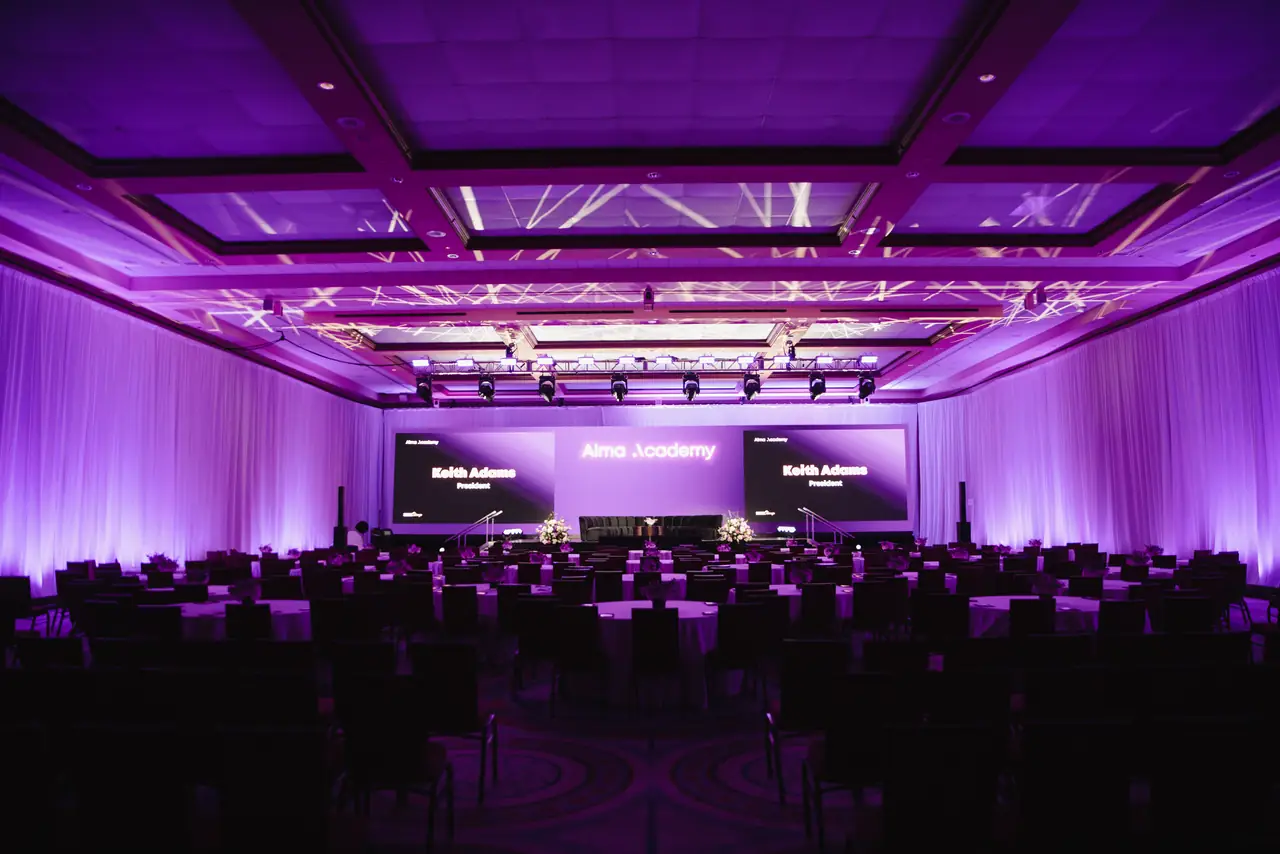 Alma Academy ballroom from the back of the room: three screens, purple drape uplight, lighting truss over rounds