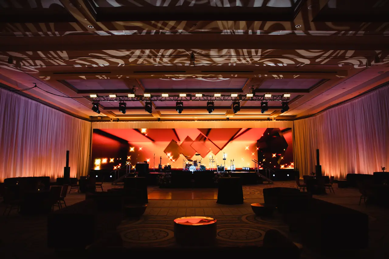 The Alma ballroom at night: warm stage light, chandeliers dimmed, rounds set for dinner